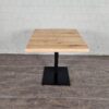 Das Foto zeigt einen minimalistischen Tisch. Die Tischplatte besteht aus hellem Holz mit einer glatten, rechteckigen Form. Sie ruht auf einem zentralen, schwarzen Metallfuß, der auf einer quadratischen Basis steht. Der Hintergrund zeigt eine weiße Ziegelwand, und der Boden besteht aus einem dunklen Holz- oder Laminatboden. Der Stil des Tisches wirkt modern und funktional, ideal für Cafés, Büros oder schlichte Wohnräume.