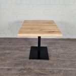 Das Foto zeigt einen minimalistischen Tisch. Die Tischplatte besteht aus hellem Holz mit einer glatten, rechteckigen Form. Sie ruht auf einem zentralen, schwarzen Metallfuß, der auf einer quadratischen Basis steht. Der Hintergrund zeigt eine weiße Ziegelwand, und der Boden besteht aus einem dunklen Holz- oder Laminatboden. Der Stil des Tisches wirkt modern und funktional, ideal für Cafés, Büros oder schlichte Wohnräume.